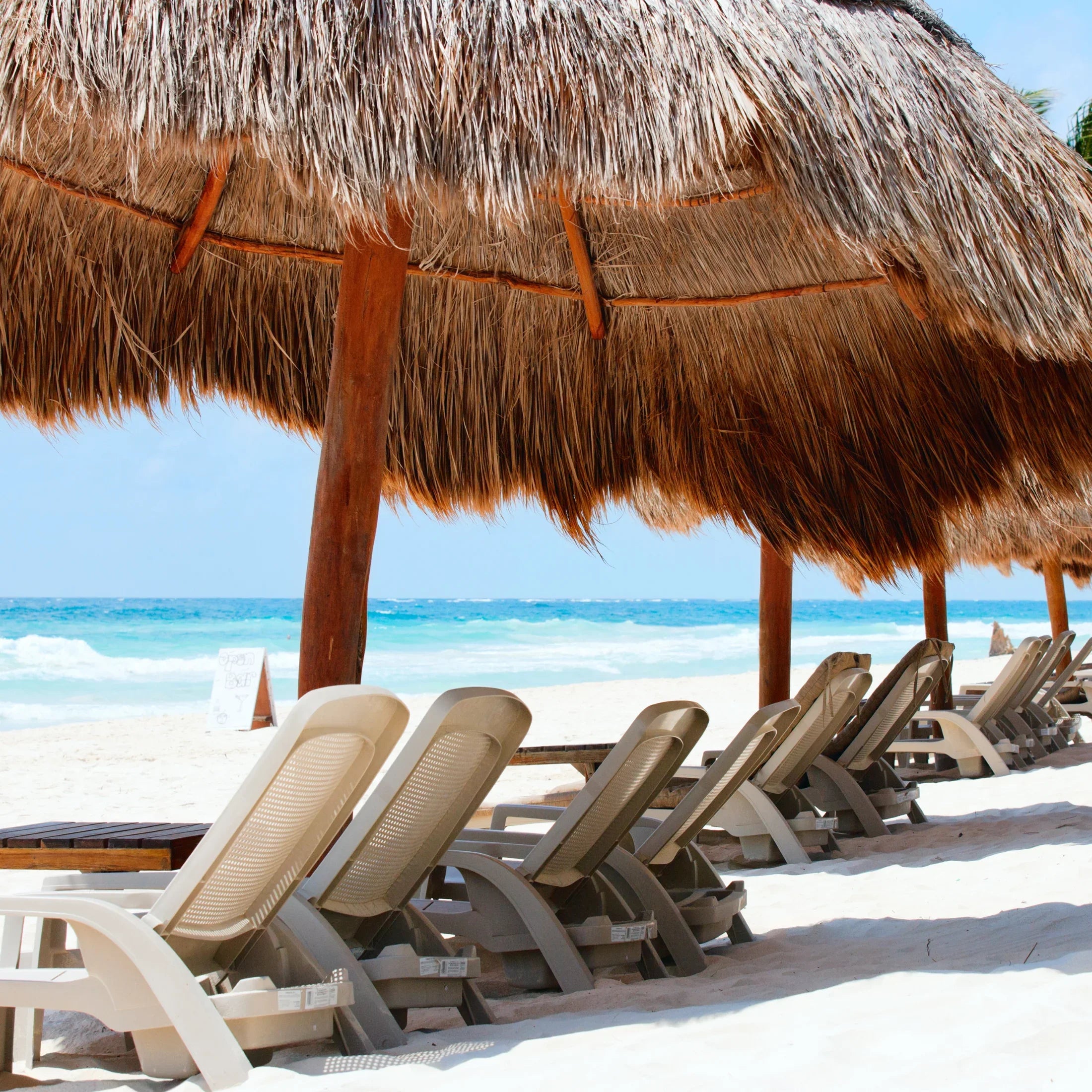 Lounge chairs under thatched beach cabanas facing ocean waves on a sunny sandy shore